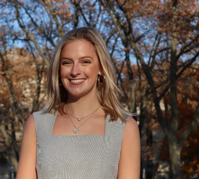 Young blonde woman smiling wearing a grey blouse with trees in the background